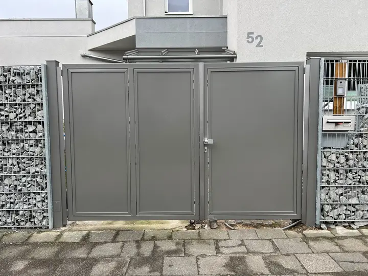 Graues Stahltor mit drei geschlossenen Blechfeldern und schmalem rechten Gehflügel zwischen Gabionen