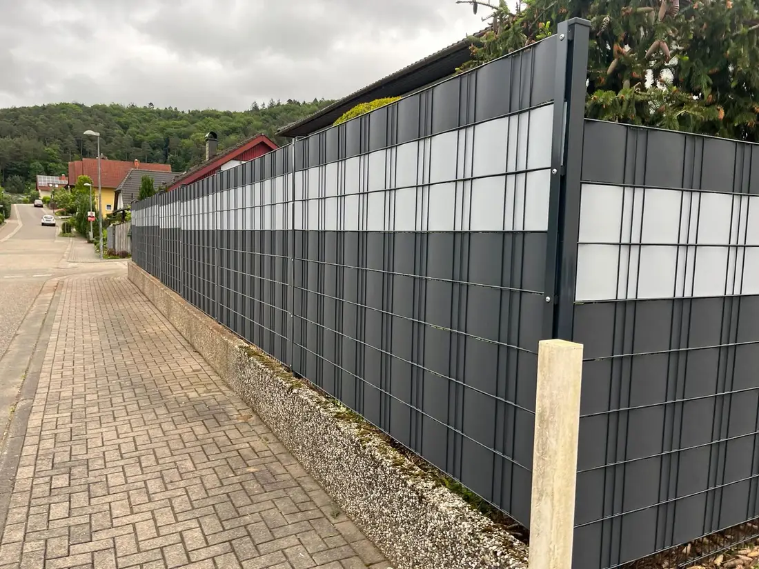 Doppelstabmattenzaun in Anthrazit mit weißen Sichtschutzstreifen auf einer Stützmauer entlang einer Ortsstraße in Waldfischbach-Burgalben