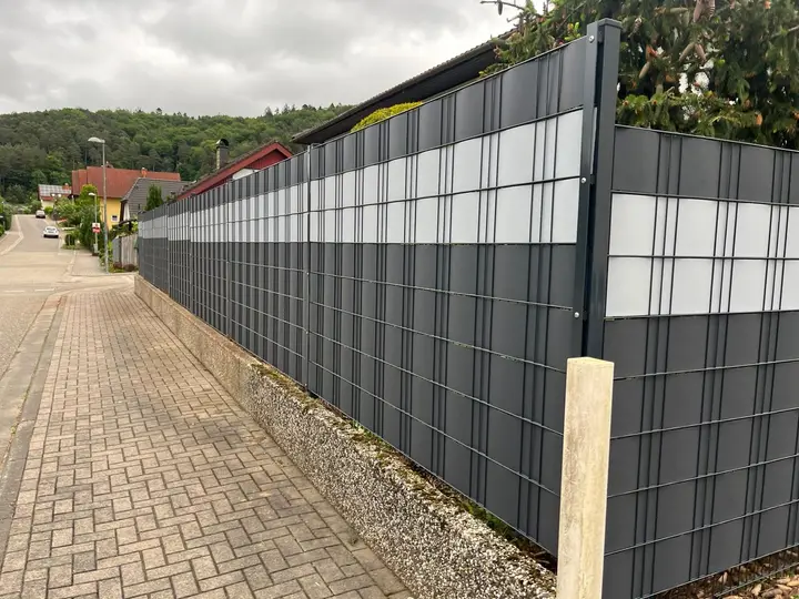 Doppelstabmattenzaun in Anthrazit mit weißen Sichtschutzstreifen auf einer Stützmauer entlang einer Ortsstraße