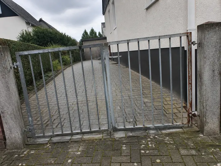 Verwittertes zweiflügeliges Metalltor mit Rost am Scharnier vor einer gepflasterten Wohnhauseinfahrt