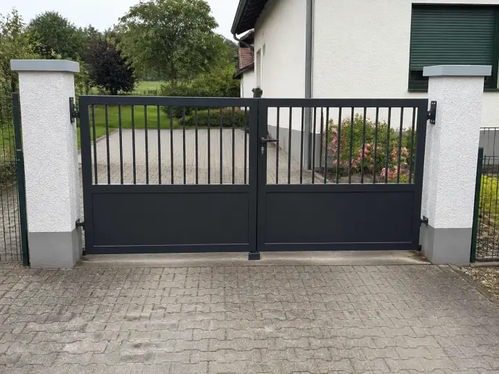 Zweiflügeliges Hoftor aus Metall in Anthrazit mit senkrechten Stäben und geschlossener unterer Füllung zwischen weißen Putzpfeilern